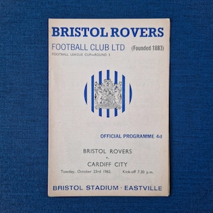 May include: A vintage Bristol Rovers Football Club programme from 23rd October 1962, advertising a match against Cardiff City. The programme features the team's crest and details about the game, including the date, time, and location.
