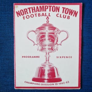 May include: A vintage Northampton Town Football Club programme cover. The cover is white with red text and a red illustration of a trophy. The text reads "Northampton Town Football Club", "Programme", "Sixpence", and "Champions Division III 1962-63".