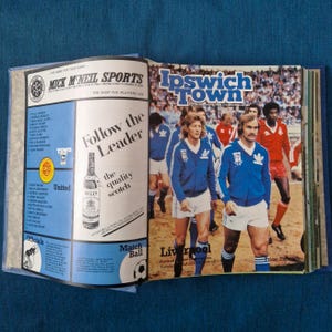 May include: A vintage Ipswich Town football match day programme featuring a blue and white cover with a photograph of players in blue track jackets and white shorts. The left page displays advertisements for Mick McNeil Sports and Bell's Scotch.