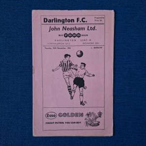 May include: A vintage pink and white programme for a Darlington F.C. football match on Tuesday, 26th December, 1961. The programme features a black and white illustration of two players in action. The programme advertises John Neasham Ltd., a Ford dealer, and Esso Golden petrol.