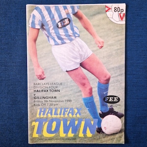 May include: A Halifax Town matchday programme from a Barclays League Division Four game. The cover shows a player in a blue and white striped shirt, white shorts, and blue socks, kicking a football. Text includes match details.