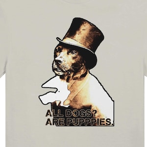May include: A light gray t-shirt featuring a dog wearing a top hat. The dog is illustrated in a sepia tone, with the text "ALL DOGS? ARE PUPPIES." printed below the dog's image. The shirt is a casual wear item.
