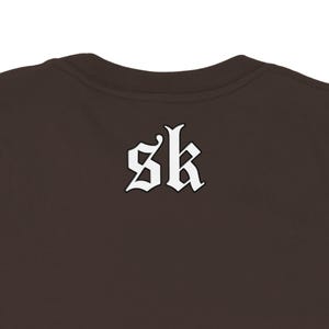 May include: A brown crew neck shirt with the white letters "sk" in a gothic font on the back. The shirt is a solid color and has a simple, classic design.