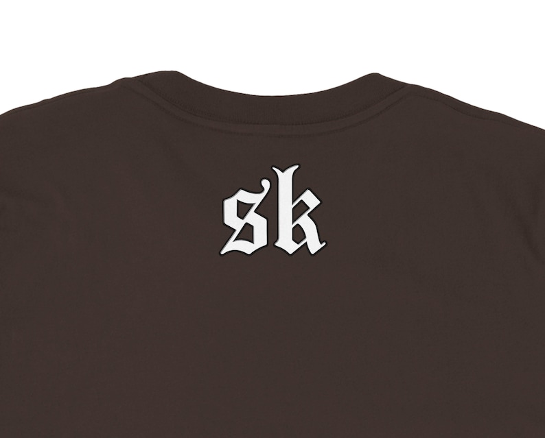 May include: A brown crew neck shirt with the white letters "sk" in a gothic font on the back. The shirt is a solid color and has a simple, classic design.