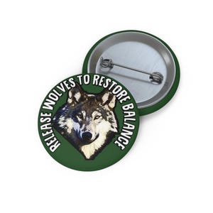 May include: A round pin-back button featuring a graphic of a wolf head centered on a dark green background. The text "RELEASE WOLVES TO RESTORE BALANCE" is printed in white capital letters in a circular arc around the wolf illustration.