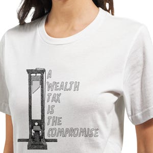 May include: White t-shirt featuring a black and white illustration of a guillotine. The text on the shirt reads "A Wealth Tax Is The Compromise". The design is centered on the front of the shirt.