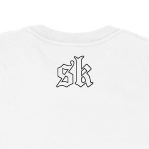 May include: White crew neck t-shirt with a black gothic-style "SK" logo on the back. The logo is outlined in black, creating a bold contrast against the white fabric. The shirt's neckline is visible.