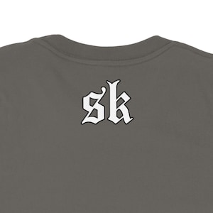 May include: A dark gray t-shirt with the white letters "sk" in a gothic font on the back. The shirt has a crew neck and is made of a soft material.