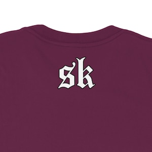 May include: Burgundy-colored t-shirt with the white letters "sk" in a gothic font on the back. The shirt's neckline is visible at the top of the frame, showcasing the fabric's texture and color.