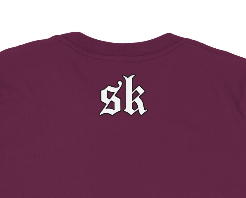 May include: Burgundy-colored t-shirt with the white letters "sk" in a gothic font on the back. The shirt's neckline is visible at the top of the frame, showcasing the fabric's texture and color.