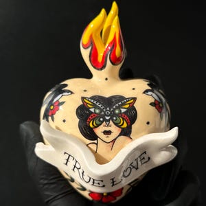 May include: Cream-colored heart-shaped ceramic sculpture with a flame design on top. The heart features a woman with a butterfly mask, floral accents, and a banner that reads "TRUE LOVE".