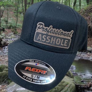 May include: A black baseball cap with a brown patch that says "Professional Asshole" in white lettering. The cap has a Flexfit label on the back.