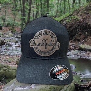May include: A black and gold baseball cap with the text "Let's keep the Dumbfuckery to a minimum today" on the front.