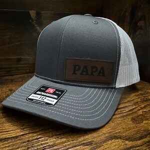 May include: A gray and white trucker hat with a brown leather patch that says "PAPA" on it. The hat is adjustable and has a Richardson 112 label.
