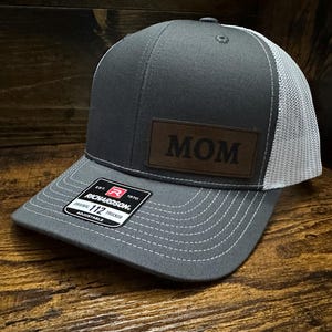 May include: A gray and white trucker hat with a brown leather patch that says "MOM" on it. The hat is adjustable and has a Richardson 112 label.