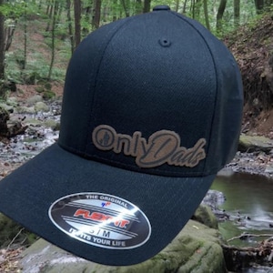 May include: Black baseball cap with a brown leather patch that says "Only Dads" in a stylized font. The cap has a Flexfit logo on the underside of the brim.