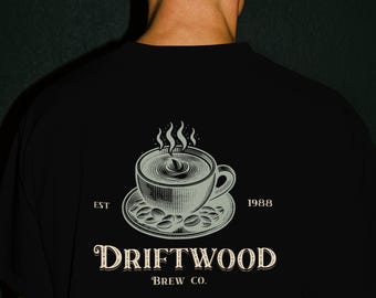 May include: Black t-shirt featuring a coffee cup design with steam, on a saucer with coffee beans. The text "DRIFTWOOD BREW CO." is below the graphic, with "EST 1988" on either side. The shirt is worn by a person.
