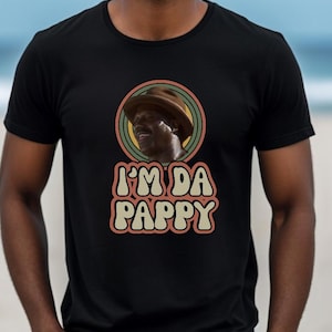 May include: Black crew neck t-shirt featuring a retro graphic of a man wearing a hat, surrounded by a circular design with green, yellow, and brown stripes. The text "I'M DA PAPPY" is below the graphic.