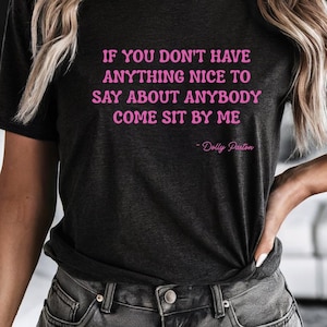 Può includere: Una t-shirt a maniche corte color grigio antracite con il testo rosa "IF YOU DON'T HAVE ANYTHING NICE TO SAY ABOUT ANYBODY COME SIT BY ME" seguito dalla firma "- Dolly Parton" stampato al centro della parte anteriore.