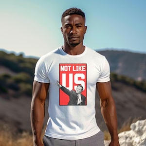 Könnte beinhalten: Weißes T-Shirt mit einer roten Grafik eines Mannes mit erhobenem Arm und dem Text "NOT LIKE US" in Weiß.