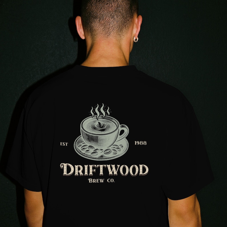 May include: Black t-shirt featuring a coffee cup design with steam, on a saucer with coffee beans. The text "DRIFTWOOD BREW CO." is below the graphic, with "EST 1988" on either side. The shirt is worn by a person.