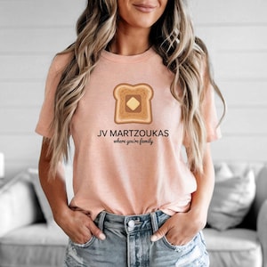 Può includere: Una maglietta color pesca con una grafica di una fetta di pane tostato con burro. Il testo "JV MARTZOUKAS where you're family" è stampato sotto la grafica del toast. La maglietta è realizzata in un materiale morbido.
