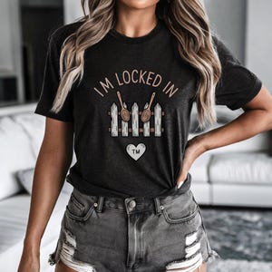 Peut inclure: T-shirt gris foncé avec l'inscription "I'M LOCKED IN" au-dessus d'un motif de clôture avec des cuillères en bois et un cœur. Le t-shirt a un col rond et des manches courtes. Le motif est dans les tons de marron et de blanc.