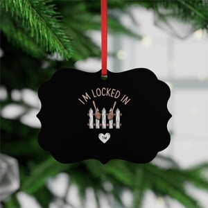 Peut inclure: Ornement de Noël en métal noir avec un bord festonné, un ruban rouge et le texte "I'M LOCKED IN". L'ornement présente une clôture blanche avec des cuillères en bois et un cœur. L'ornement est suspendu devant un sapin de Noël vert.