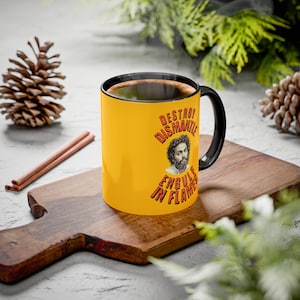 Peut inclure: Une tasse à café jaune avec une anse et un bord noirs. La tasse présente une image d'un homme et les mots "DESTROY, DISMANTLE, ENGULF IN FLAMES" en rouge et orange. La tasse est sur une planche à découper en bois.