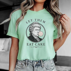 May include: A pale green t-shirt with the words "LET THEM EAT CAKE" printed in black. The graphic is a portrait of a historical figure blowing a bubblegum bubble. The shirt is made of a soft material.