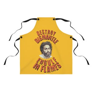 May include: Mustard yellow apron with black ties. The apron features a portrait of a man with curly hair and the words "DESTROY DISMANTLE ENGULF IN FLAMES" in red letters.