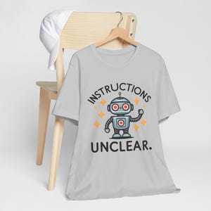 以下が含まれることがあります： ロボットのグラフィックと「INSTRUCTIONS UNCLEAR.」というテキストが書かれた薄いグレーのTシャツ