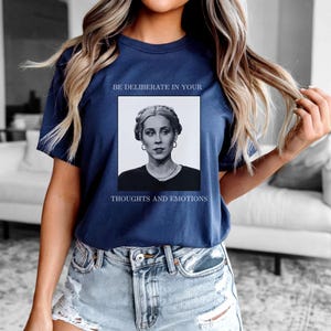 May include: Navy blue t-shirt featuring a black and white portrait of a woman with the text "BE DELIBERATE IN YOUR THOUGHTS AND EMOTIONS". The shirt is a classic crew neck style.