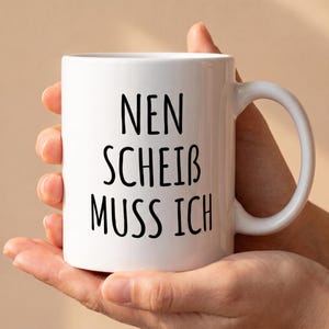 Può includere: Una tazza in ceramica bianca con testo nero stampato sul davanti che recita NEN SCHEIB MUSS ICH in un semplice carattere sans-serif. La tazza presenta una finitura liscia e lucida e un manico curvo sul lato destro per le bevande calde.