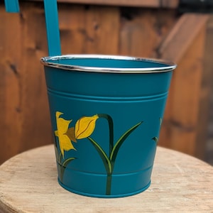 Könnte beinhalten: Türkisfarbener Metall-Hängepflanztopf mit silbernem Rand und türkisfarbenem Griff. Der Pflanztopf hat ein handgemaltes gelbes Narzissen-Design mit grünen Blättern. Der Pflanztopf steht auf einem Holzschemel.