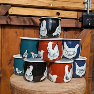 May include: A selection of enamel mugs in colours such as black, teal, red, and blue. Each mug is decorated with a white chicken design. The mugs are stacked on a wooden stool, set against a wooden door.