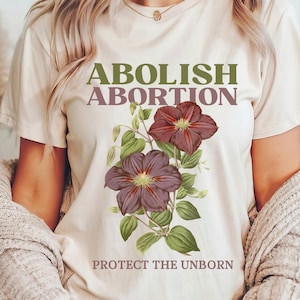 Puede incluir: Camiseta color crema con el texto "ABOLISH ABORTION" en verde y morado, con "PROTECT THE UNBORN" debajo. La camiseta muestra un diseño floral con dos flores granates y hojas verdes.