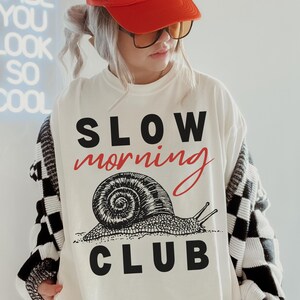 K&ouml;nnte beinhalten: Wei&szlig;es T-Shirt mit der Aufschrift "SLOW morning CLUB" und einer Schnecken-Grafik. Die Person tr&auml;gt eine rote Trucker-M&uuml;tze mit der Aufschrift "MAD LOVE FOR YOU". Sie tr&auml;gt auch eine Brille und eine schwarz-wei&szlig; karierte Strickjacke.