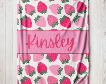Puede incluir: Una manta personalizada rosa y blanca con un patrón de fresas. El nombre "Kinsley" está impreso en rosa sobre una pancarta rosa.