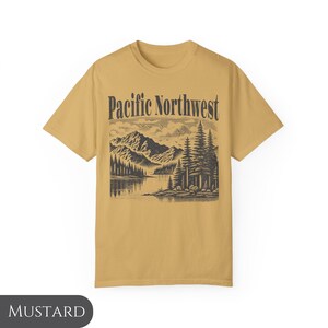 Może przedstawiać: Ż&oacute;łta koszulka w kolorze musztardowym z czarno-białym nadrukiem graficznym g&oacute;rskiego krajobrazu i tekstem "Pacific Northwest".