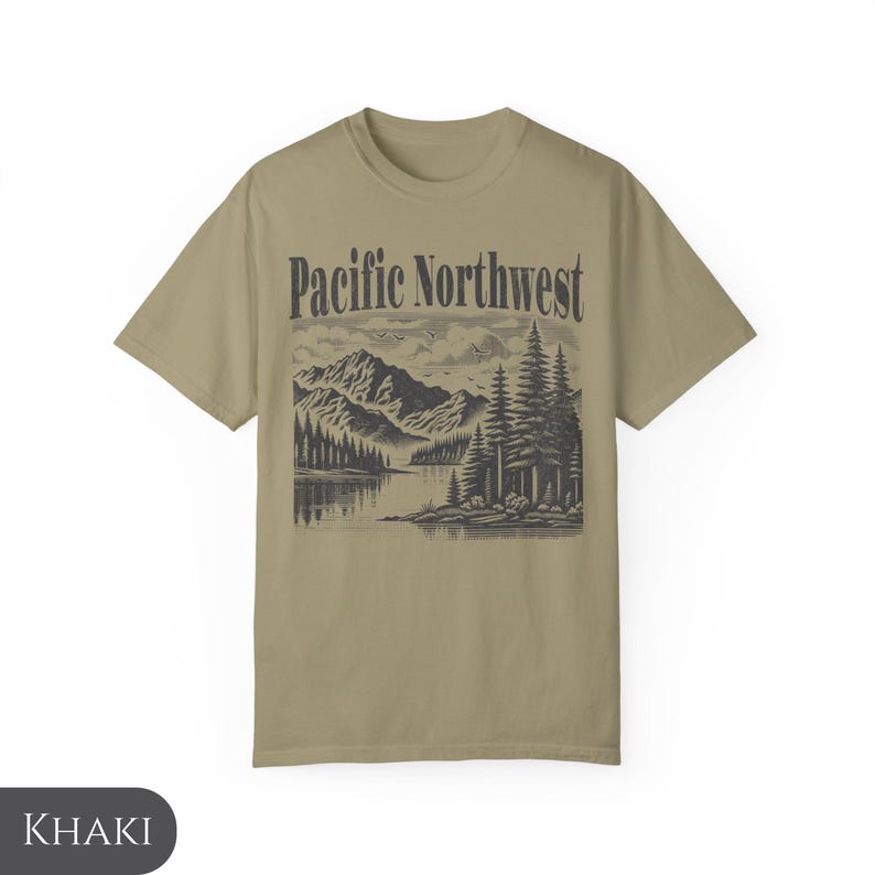 Może przedstawiać: Koszulka w kolorze khaki z czarno-białym nadrukiem przedstawiającym pasmo g&oacute;rskie, jezioro i drzewa. Tekst "Pacific Northwest" jest nadrukowany na czarno nad grafiką.