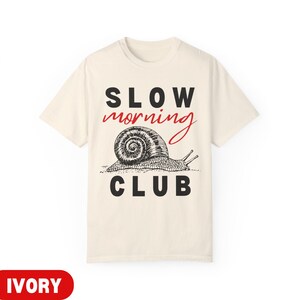 K&ouml;nnte beinhalten: Elfenbeinfarbenes T-Shirt mit dem Schriftzug "SLOW MORNING CLUB" in Schwarz und Rot. Darunter befindet sich eine detaillierte Schwarz-Wei&szlig;-Illustration einer Schnecke. Das T-Shirt ist ein Freizeitartikel.