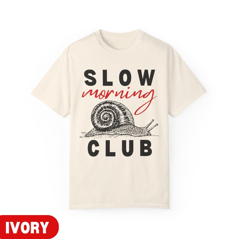 K&ouml;nnte beinhalten: Elfenbeinfarbenes T-Shirt mit dem Schriftzug "SLOW MORNING CLUB" in Schwarz und Rot. Darunter befindet sich eine detaillierte Schwarz-Wei&szlig;-Illustration einer Schnecke. Das T-Shirt ist ein Freizeitartikel.