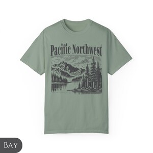 Może przedstawiać: Zielony t-shirt w kolorze szałwii z czarno-białym nadrukiem graficznym przedstawiającym pasmo g&oacute;rskie, jezioro i drzewa. Tekst "Pacific Northwest" jest nadrukowany czarnym kolorem nad grafiką.