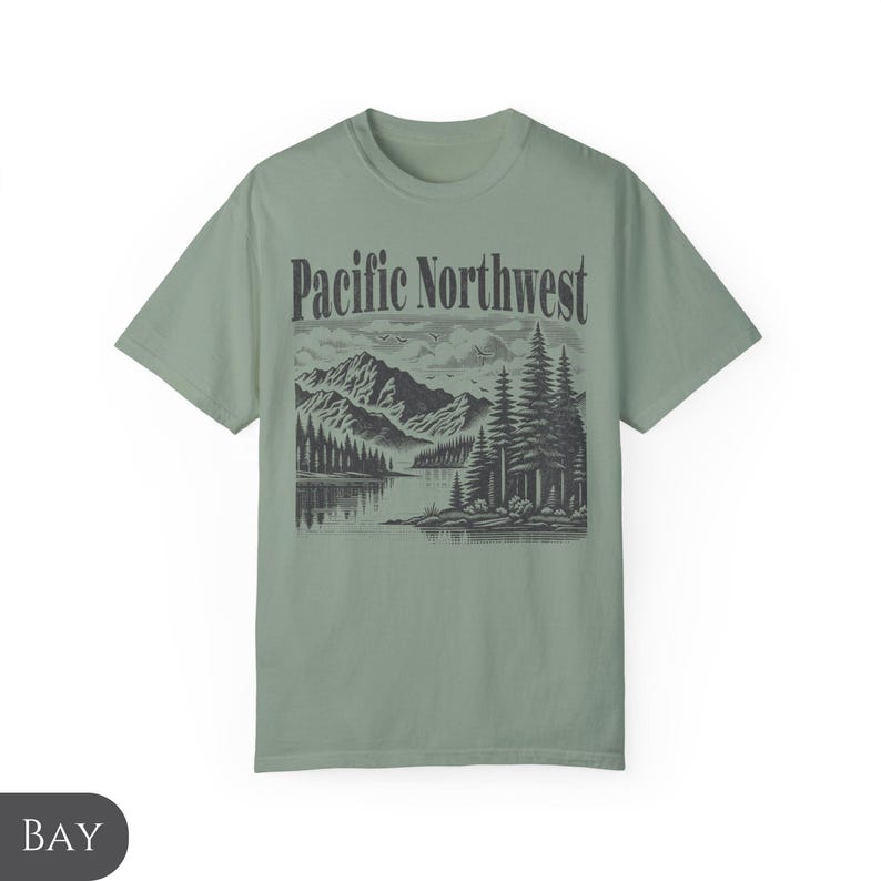 Może przedstawiać: Zielony t-shirt w kolorze szałwii z czarno-białym nadrukiem graficznym przedstawiającym pasmo g&oacute;rskie, jezioro i drzewa. Tekst "Pacific Northwest" jest nadrukowany czarnym kolorem nad grafiką.