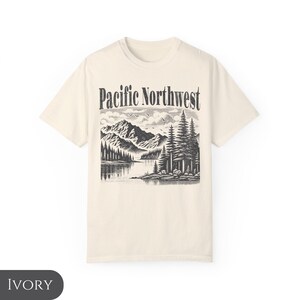 Może przedstawiać: Kremowa koszulka z czarno-białym nadrukiem graficznym przedstawiającym pasmo g&oacute;rskie i drzewa. Tekst "Pacific Northwest" jest nadrukowany na g&oacute;rze grafiki czarnym kolorem.