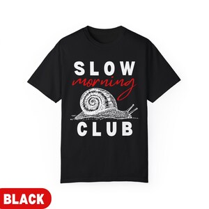 K&ouml;nnte beinhalten: Schwarzes T-Shirt mit dem Schriftzug "SLOW MORNING CLUB" in Wei&szlig; und Rot. Unter dem Text befindet sich eine detaillierte Illustration einer Schnecke. Das T-Shirt hat einen Rundhalsausschnitt.