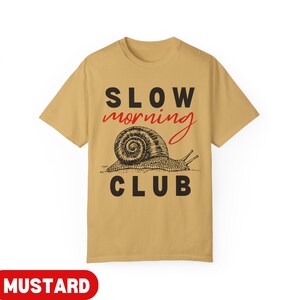 K&ouml;nnte beinhalten: Senffarbenes T-Shirt mit dem Schriftzug "SLOW morning CLUB" in Schwarz und Rot. Unter dem Text befindet sich eine detaillierte Illustration einer Schnecke. Dieses T-Shirt ist ein l&auml;ssiges Kleidungsst&uuml;ck.