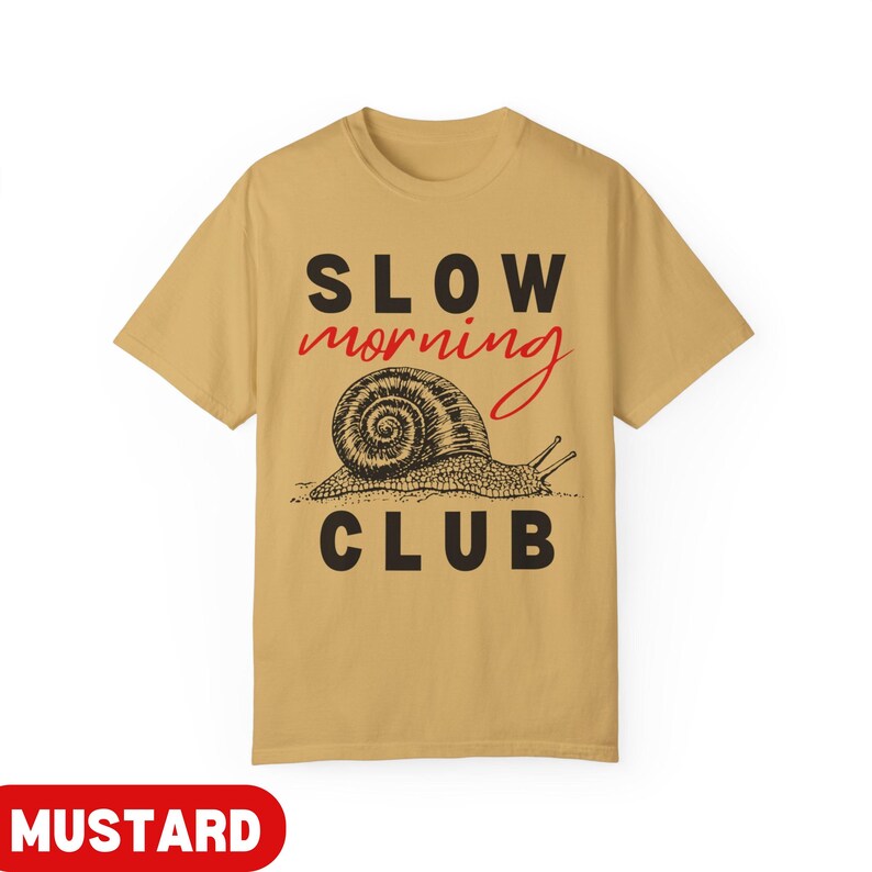K&ouml;nnte beinhalten: Senffarbenes T-Shirt mit dem Schriftzug "SLOW morning CLUB" in Schwarz und Rot. Unter dem Text befindet sich eine detaillierte Illustration einer Schnecke. Dieses T-Shirt ist ein l&auml;ssiges Kleidungsst&uuml;ck.