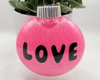 May include: A pink glitter Christmas ornament with the word "LOVE" in black block letters. The ornament has a silver metal cap and a silver ribbon for hanging. A green plant in a white pot is in the background.